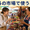 海外の市場で商品を手に取り、店員に英語で値段を尋ねる日本人旅行者