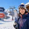 go skiing、lift ticket、ski rentalの看板があるスキー場でスキーを持つ女性